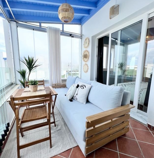 A bright sunlit balcony features a large, cozy sofa complemented by decorative cushions. A wooden table and two chairs are positioned nearby, creating a comfortable dining space. Large windows provide a panoramic view, and light curtains allow for gentle natural illumination.