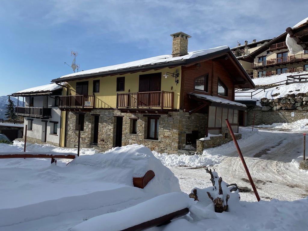 A chalet-style building is presented, featuring a combination of stone and wood siding. Snow covers the ground, highlighting the winter setting. Balconies with wooden railings are visible on the upper levels, and large windows allow natural light to enter. The surrounding landscape includes snow-laden pathways.