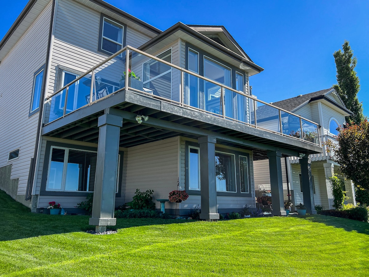 The exterior of the house showcases a spacious walkout basement suite, highlighted by large windows and a wrap-around balcony. A well-maintained lawn surrounds the property, with vibrant greenery visible along the base. The structure's modern design features a combination of light siding and sturdy dark pillars.