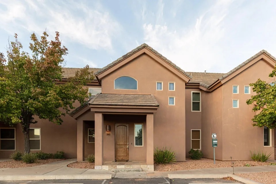 The exterior of a two-story condo is showcased, featuring adobe-style architecture with a warm, earthy color palette. The entrance is framed by a covered porch and a wooden door, while large windows reflect the surrounding greenery and desert landscaping.