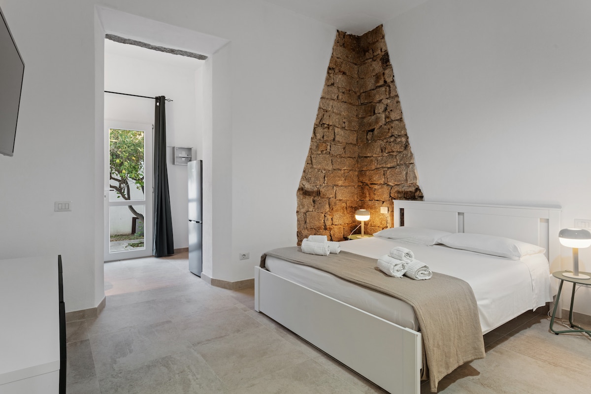 A cozy sleeping area features a spacious bed with fresh linens and a neutral-colored throw. Exposed stone walls provide texture, while a small desk and lamp enhance functionality. A doorway leads to a private garden, with natural light brightening the room through nearby windows.