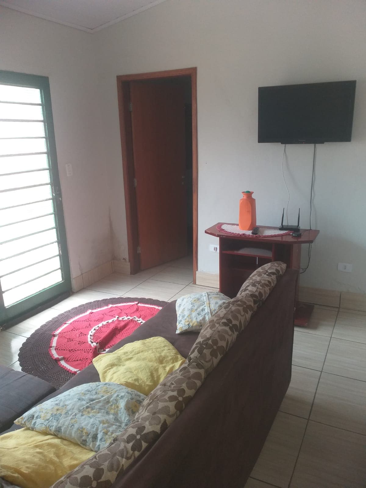 A simple living area is visible, featuring a brown sofa with colorful cushions. A television is mounted on the wall opposite a small table with a bottle. Natural light enters through a glass door, revealing a welcoming entrance.
