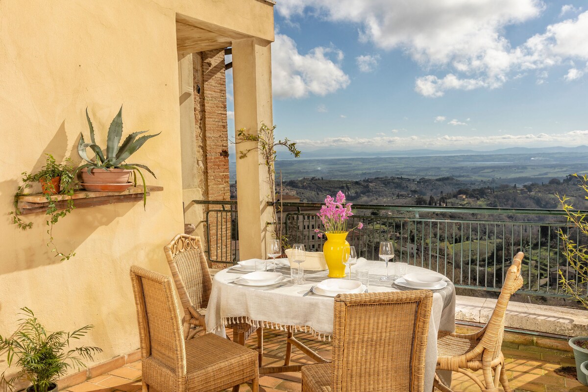 A dining table set for four is positioned on a sunny terrace, surrounded by woven chairs. A vibrant yellow vase holds fresh flowers, while scenic views of rolling hills and vineyards stretch out into the distance under a partly cloudy sky.