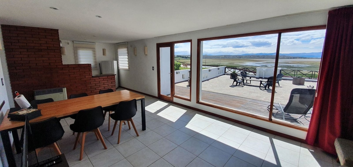 A spacious living area features a dining table with black chairs and warm wooden accents. Large windows showcase sweeping views of the landscape and outdoor seating on the terrace. Natural light brightens the room, highlighting its modern design.