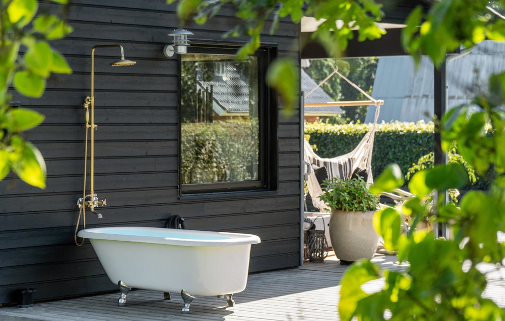 An outdoor space showcases a white freestanding bathtub, complemented by a gold shower attachment. A large window offers a view into the home, while lush green foliage frames the area, creating a serene and inviting environment for relaxation.