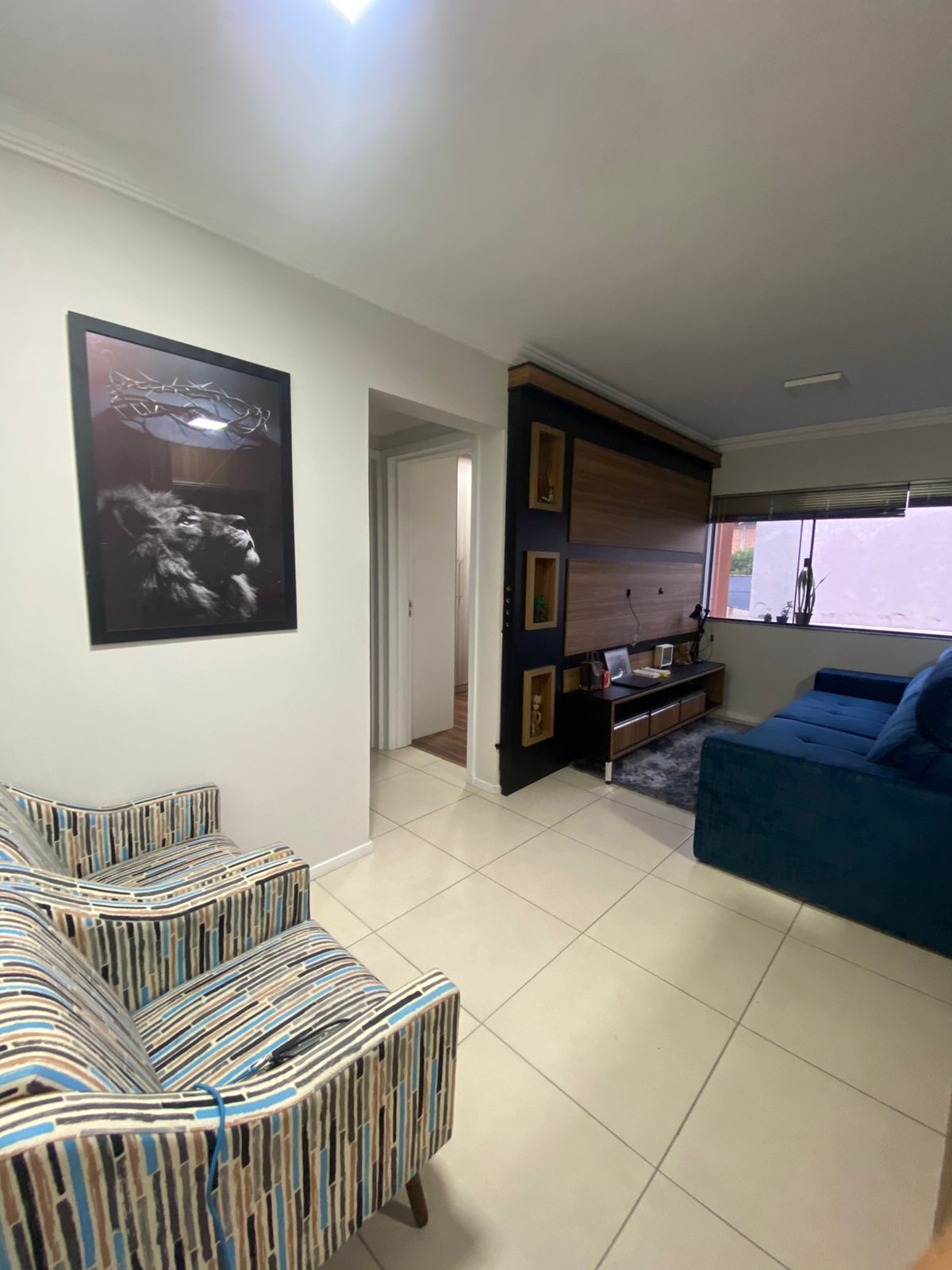 A living area is presented with striped armchairs, adding a touch of color. A wall-mounted entertainment unit complements the blue sofa. Natural light filters in through a window, creating a bright and airy space. A tasteful artwork featuring a lion decorates the wall.