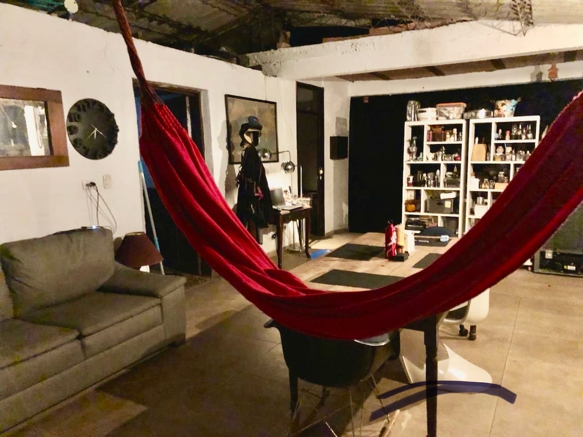 A common area is depicted, featuring a red hammock suspended between two points. A neutral-colored couch is positioned to one side, while a dining table with chairs occupies the center. Shelves filled with items are visible against the wall, enhancing the relaxed atmosphere.