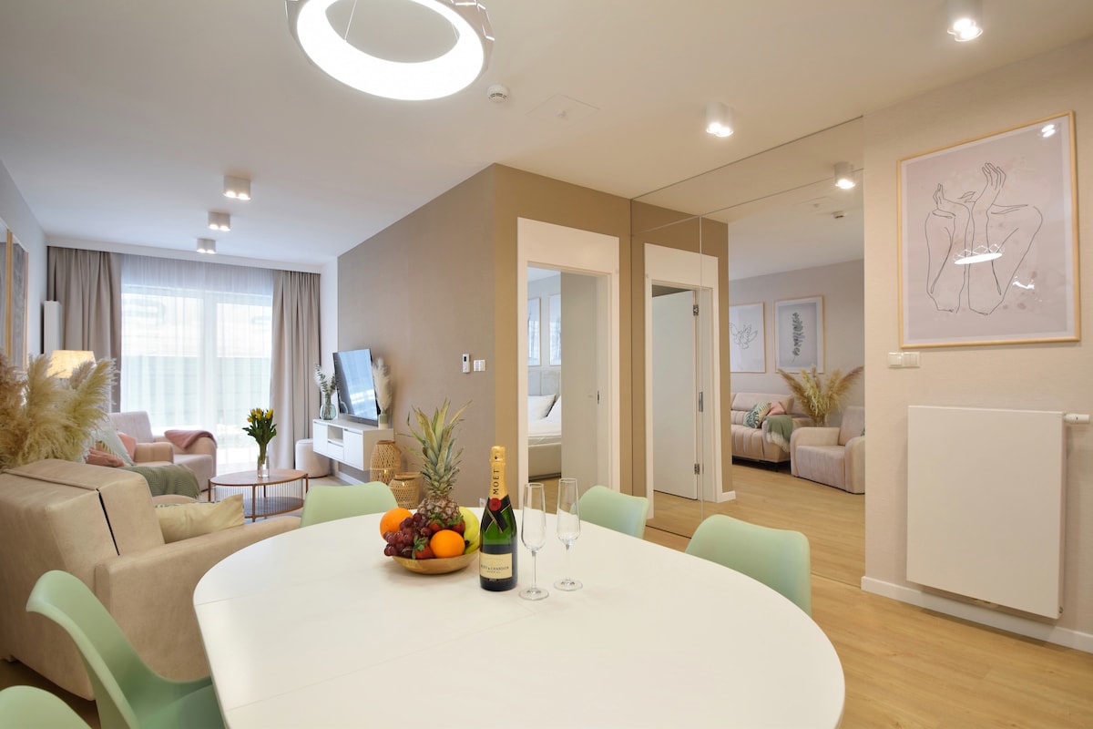 A modern dining area features a round white table surrounded by pastel green chairs, with a bottle of champagne and glasses set on the table. The open layout leads to a cozy living space, illuminated by natural light from large windows, and a subtle decorative touch is provided by wall art.