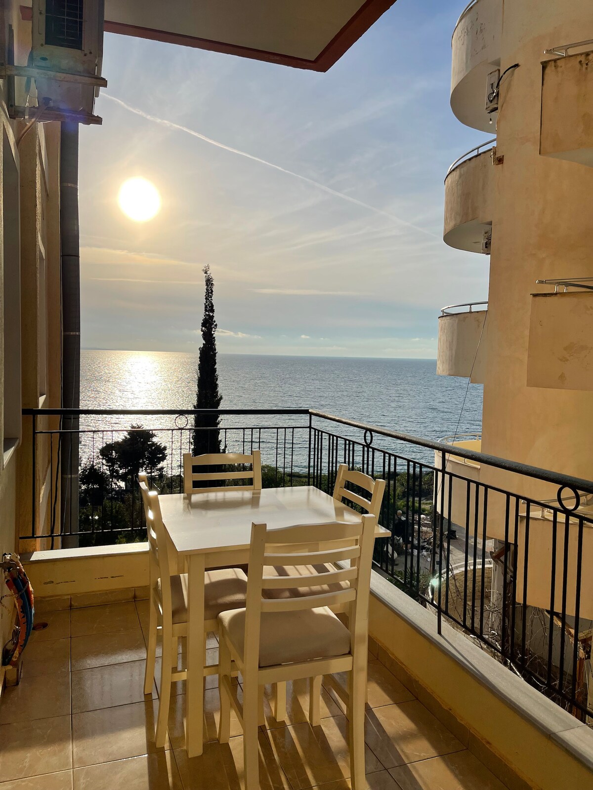 A balcony offers a dining table with four white chairs, overlooking a serene ocean view. The sun hangs low in the sky, reflecting on the water, while a tall tree emerges from the landscape, providing a natural focal point against the backdrop of the sea.