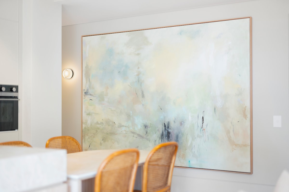 A large abstract artwork adorns the wall, featuring soft hues of blue and cream that create a calming focal point. The art is complemented by light from a nearby modern fixture, while a dining table with woven chairs is positioned in the foreground.