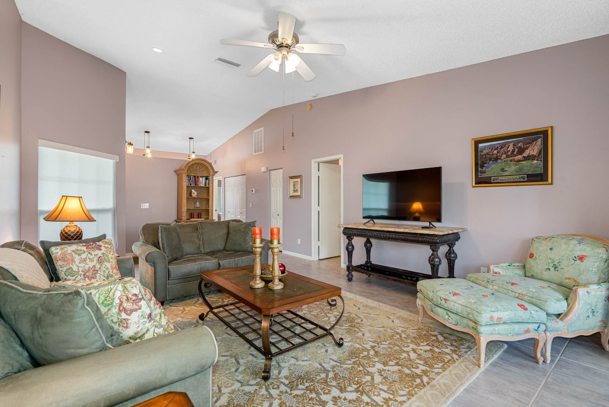 A spacious living area features comfortable seating with a blend of traditional and modern styles. A television is mounted on the wall, while a decorative rug anchors the seating arrangement. Soft lighting enhances the welcoming ambiance, complemented by natural light from nearby windows.