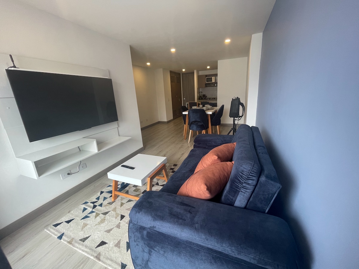 A modern living area is featured, showcasing a navy blue sofa with cushions. A flat-screen television is mounted on the wall, opposite a small coffee table situated on a patterned rug. A dining area with chairs is visible in the background.