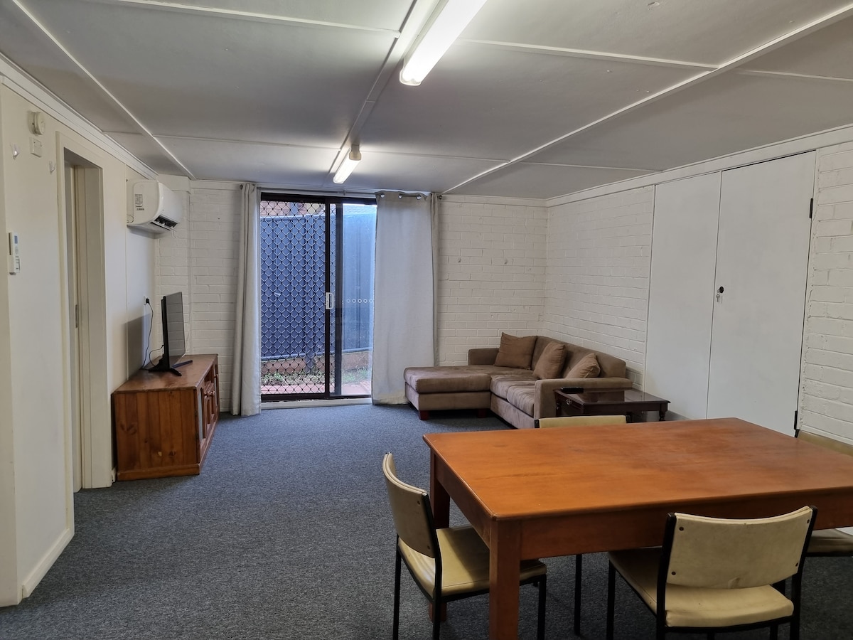 A spacious living area features a brown sectional sofa and a wooden dining table surrounded by chairs. A television is placed on a wooden cabinet, with natural light filtering through exterior doors leading to a fenced outdoor space.