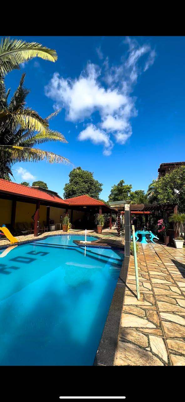 A well-maintained outdoor pool area is surrounded by lush greenery and palm trees. The pool is bordered by a stone patio, featuring colorful loungers and shaded spaces. Clear blue skies complement the vibrant atmosphere, inviting relaxation in a spacious setting.