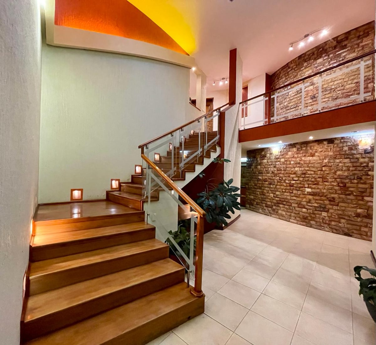 A staircase with polished wooden steps leads to an upper level, illuminated by soft lights along the side. The walls feature a combination of smooth white and rustic stone, enhancing the inviting design. Green plants are positioned at the base, complementing the layout.