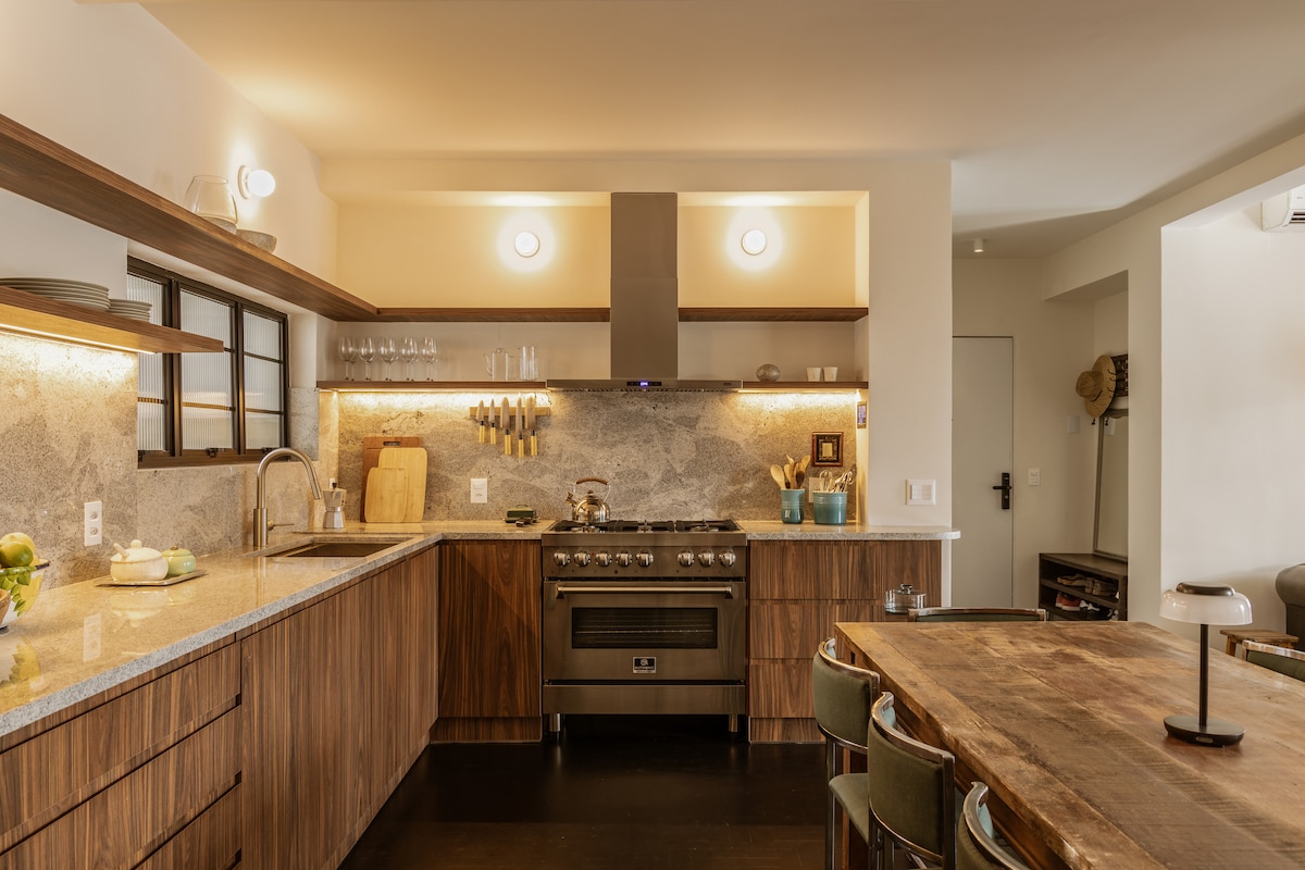 A modern kitchen with a spacious layout features sleek wooden cabinetry and a striking stone backsplash. A high-end stove is prominently displayed, accompanied by an array of cooking utensils. The dining table, crafted from wood, is set for a gathering, surrounded by comfortable chairs.