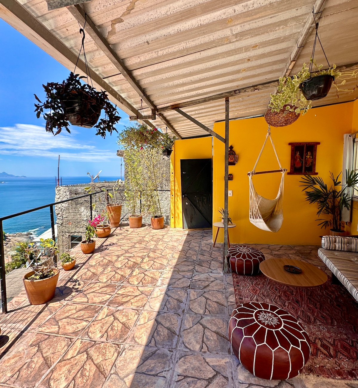 A vibrant outdoor terrace features a yellow wall and a variety of potted plants, creating a serene atmosphere. A hanging hammock is positioned near the open door, accompanied by a low circular table and traditional poufs on the tiled floor, all with views of the ocean.