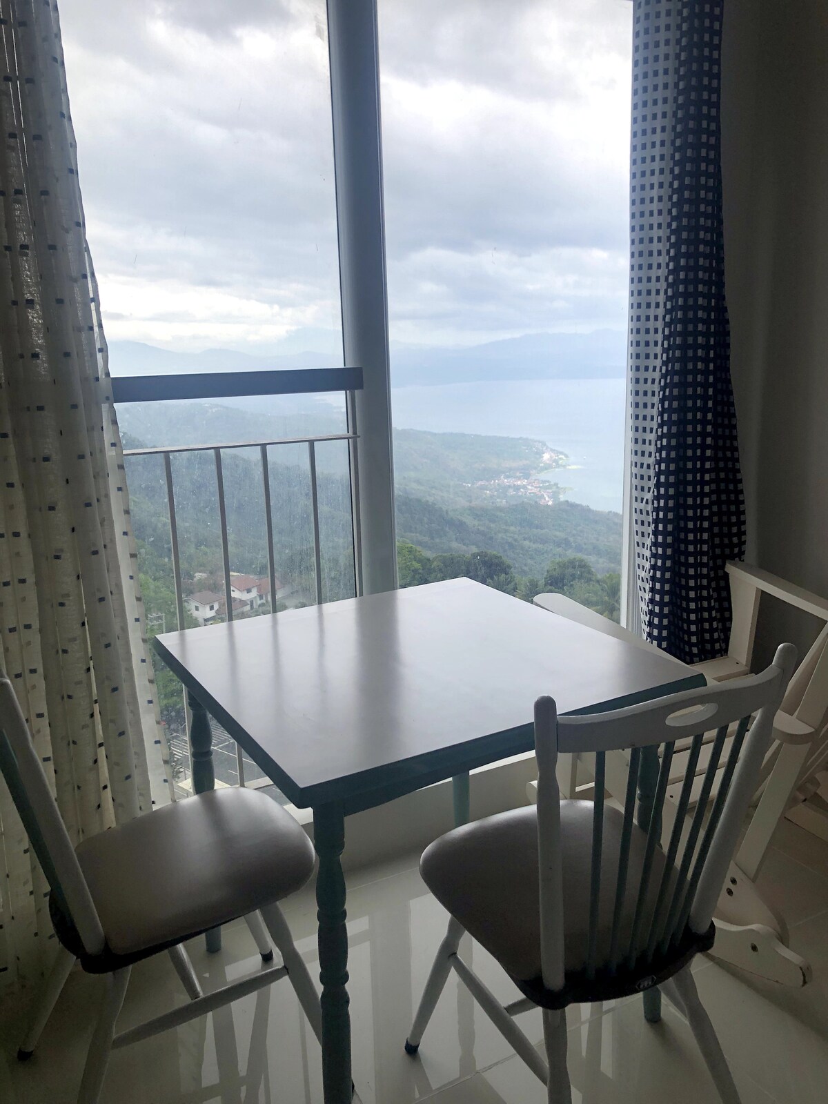 A small dining table is positioned beside a large window, offering expansive views of the surrounding landscape and ocean. Two chairs are arranged at the table. Sheer curtains frame the window, softly diffusing natural light into the space.
