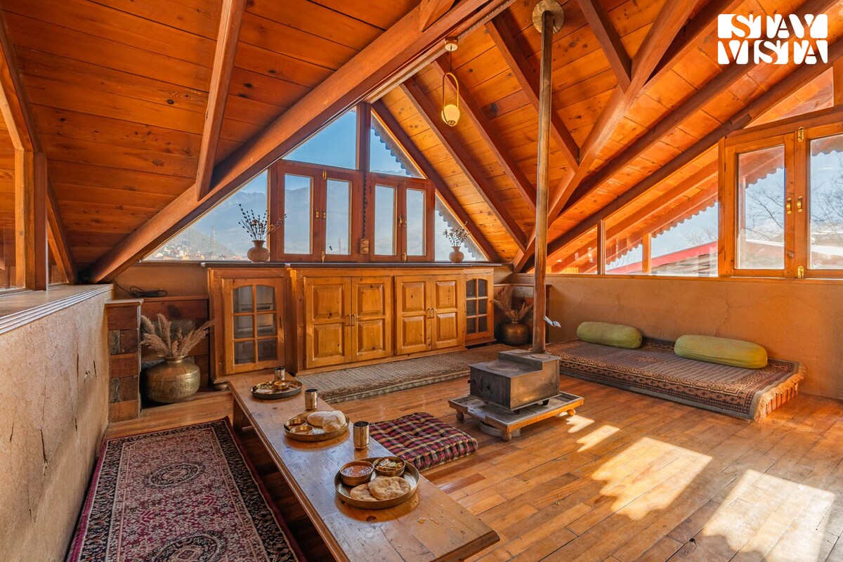 A spacious loft area features a wooden ceiling with exposed beams and large windows that allow natural light to fill the space. A low wooden table is set for dining, surrounded by cushions for seating. An ornate stove is positioned in the center for warmth.