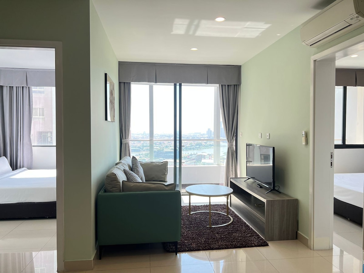 The living area features a comfortable sofa and a coffee table, with a large window that provides views of the city. Adjacent rooms with spacious beds can be seen through an open doorway, contributing to an airy feel throughout the space.