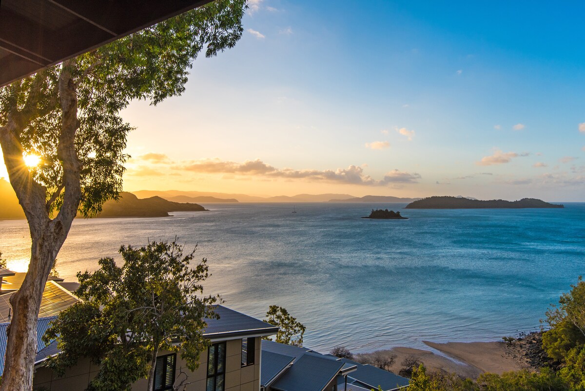 A serene coastal view captures gentle waves lapping at the shore under a soft sunset sky. Distant islands dot the horizon, framed by lush greenery. The scene reflects a tranquil atmosphere, ideal for relaxation and appreciation of the surrounding natural beauty.