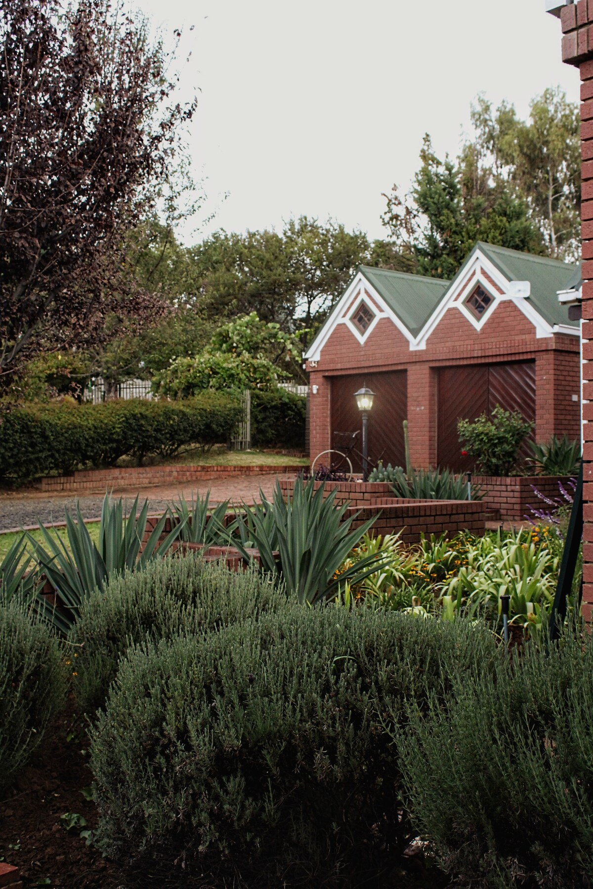 A well-maintained garden features various plants and shrubs, flanking a path leading to a red-brick house with a green roof. Decorative architectural details adorn the gables, while a lantern adds a charming touch to the outdoor space.