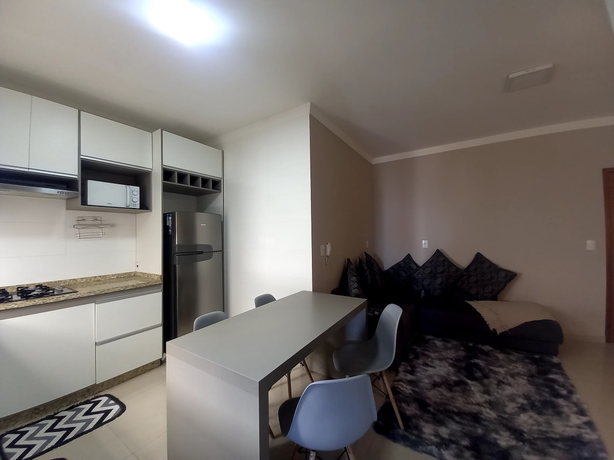 A modern kitchen area includes sleek cabinetry and essential appliances such as a cooktop, microwave, and refrigerator. A dining table with four chairs is positioned near a cozy seating area, which features a large, soft rug and floor cushions for relaxation.