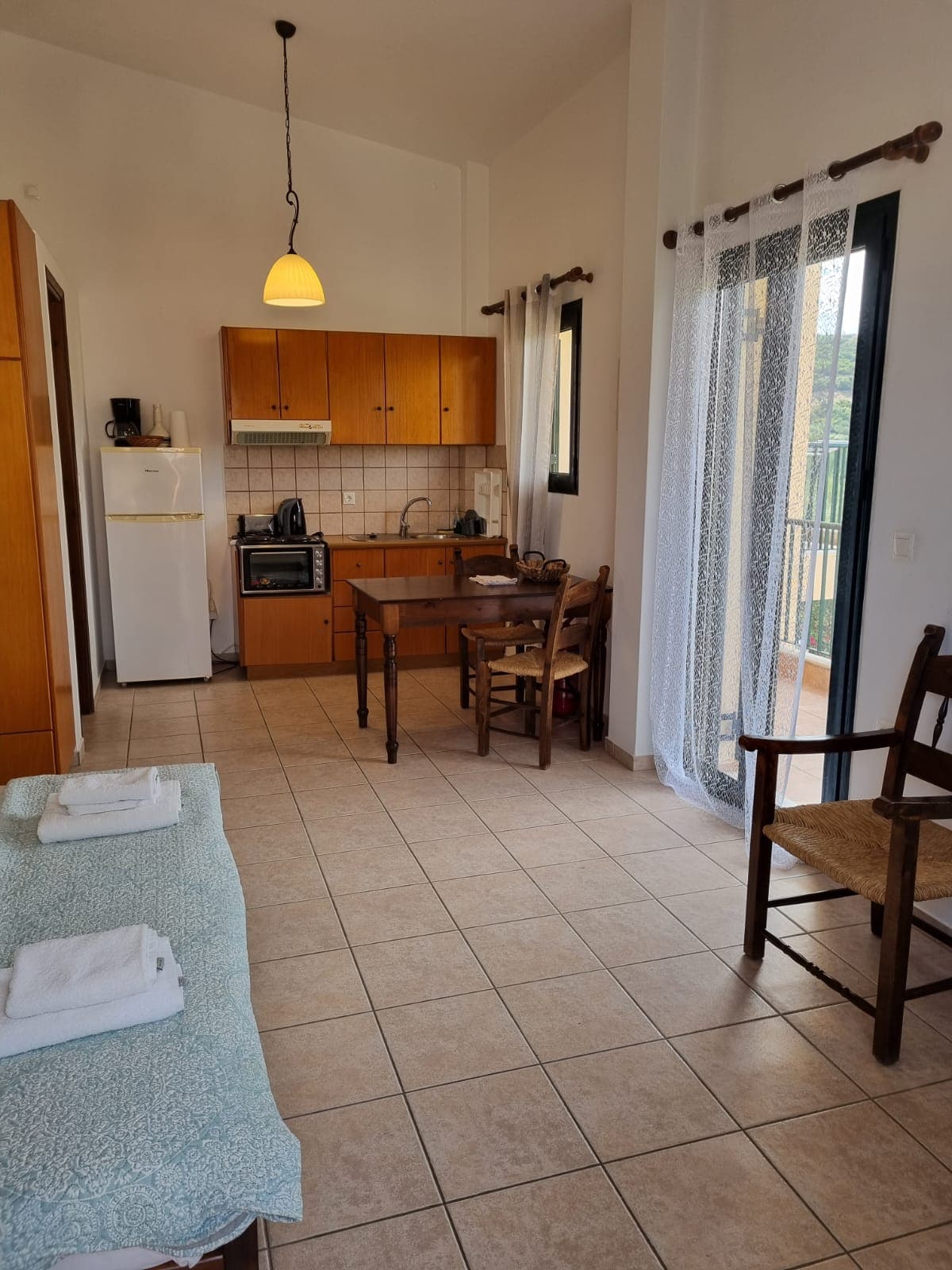 The apartment features a functional kitchenette equipped with a mini oven and refrigerator. A dining table with chairs is positioned beside the window, allowing natural light to fill the space. An inviting seating area includes a spacious couch, while light-colored tiles and soft curtains enhance the ambiance.