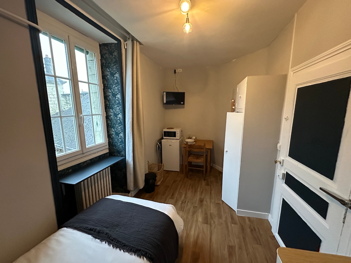 A cozy room is presented with a large window allowing natural light. A wall-mounted television is visible, along with a compact kitchenette featuring a microwave and refrigerator. A small dining table with chairs occupies a corner, adjacent to a neatly made bed with a dark blanket.