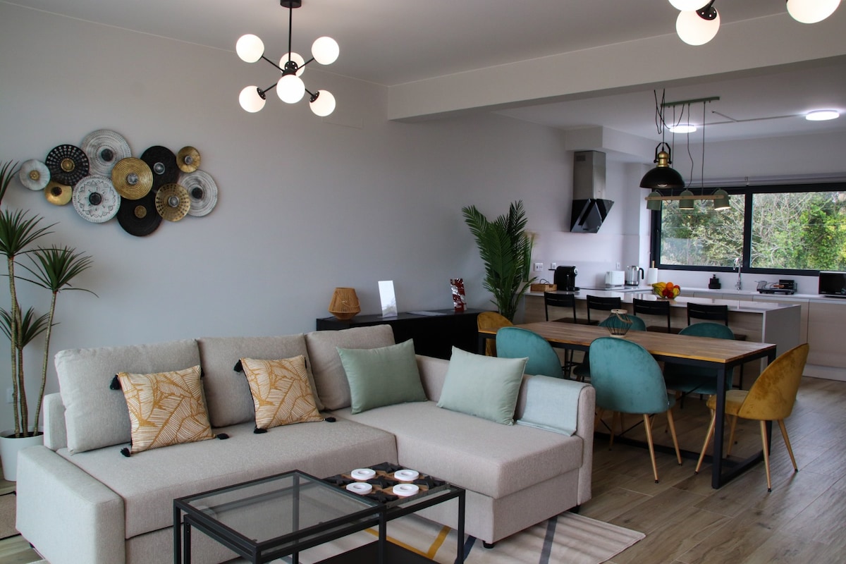 The living area features a light-colored sectional sofa adorned with colorful cushions, placed adjacent to a glass coffee table. A dining table with colorful chairs is visible in the background, complemented by modern lighting fixtures. A subtle plant adds a touch of greenery to the space.