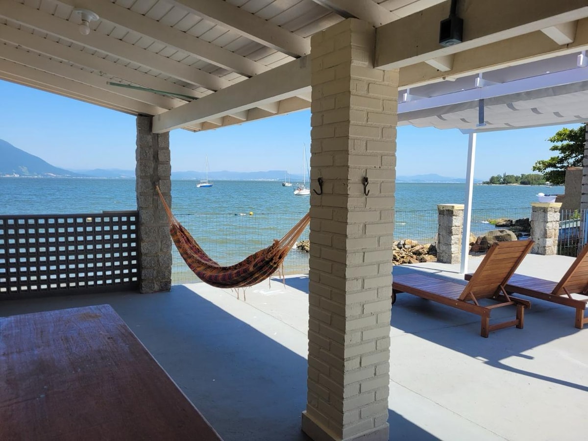 An outdoor seating area features wooden lounge chairs positioned beside a calm bay. A hammock is gently draped between two columns, while the expansive water view stretches into the distance. Sailboats can be seen bobbing quietly in the bay, framed by clear skies.