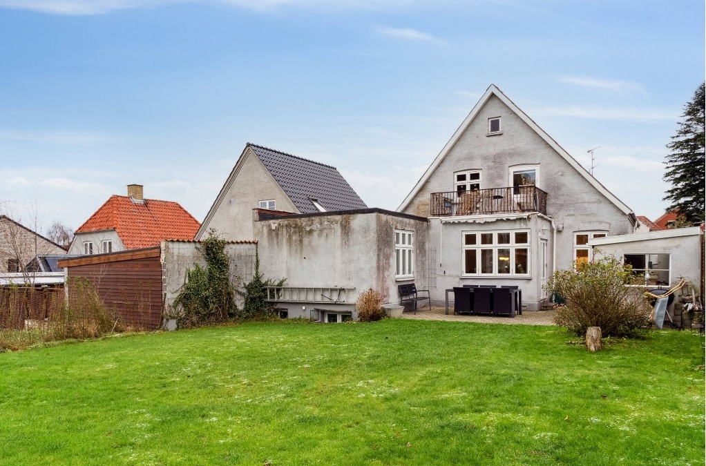 The exterior of a charming villa is presented, featuring a spacious green lawn and a mix of trees and shrubs. The back of the house is visible, showcasing large windows and a balcony. Outdoor seating is arranged on a small patio area.