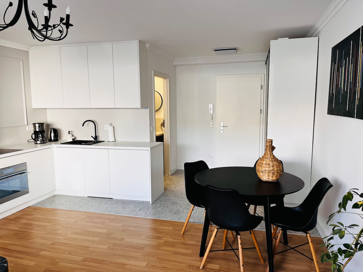 A modern kitchen features white cabinetry and a black countertop. A dining area with a round table and black chairs is seen. A woven decorative vase sits on the table. The space is completed with a doorway leading to an area beyond, complemented by a wall art piece with abstract designs.