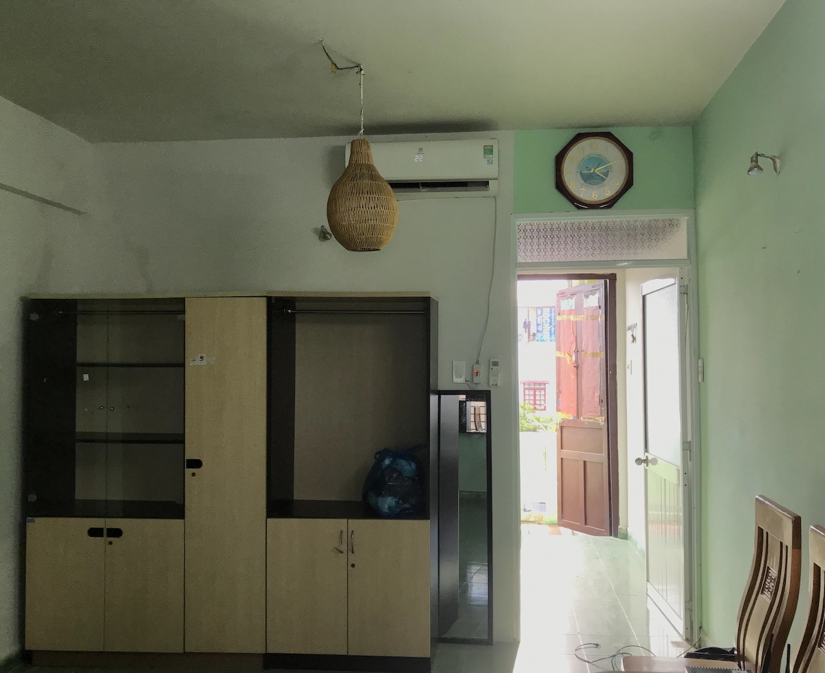 The living space features light green walls and a ceiling with a woven pendant light. A simple wooden cabinet stands against the wall with a television or decorative items. A doorway leads to an outdoor area, allowing natural light to enter the room.