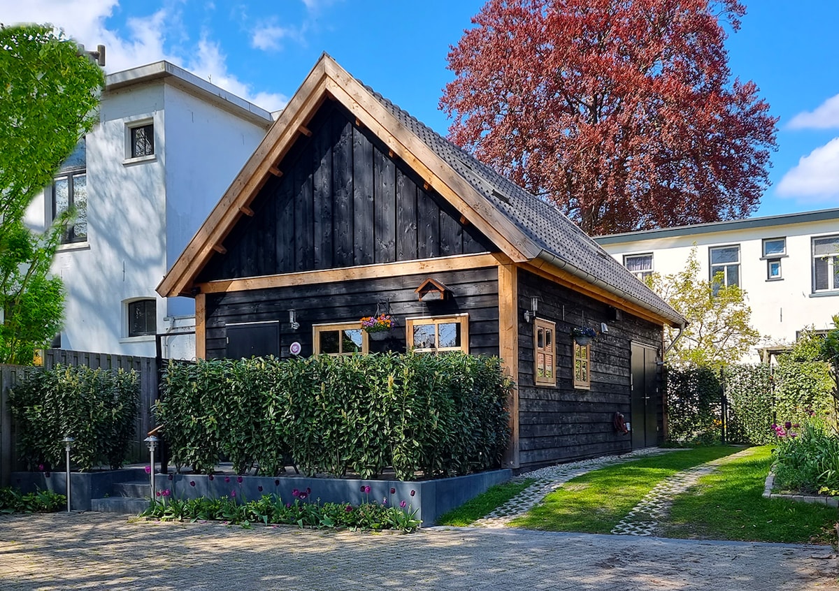 A charming black wooden house is surrounded by lush greenery and flowering plants. The roof features a gentle slope, while large windows allow light to enter. A driveway leads to the home, bordered by well-maintained grass and a pathway made of stones.