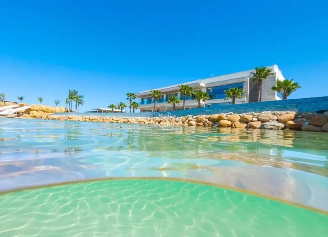 The water's surface reflects the clear blue sky, while a modern building with large windows is positioned in the background. The landscape features palm trees and smooth rocks along the water's edge, providing a serene and inviting view.
