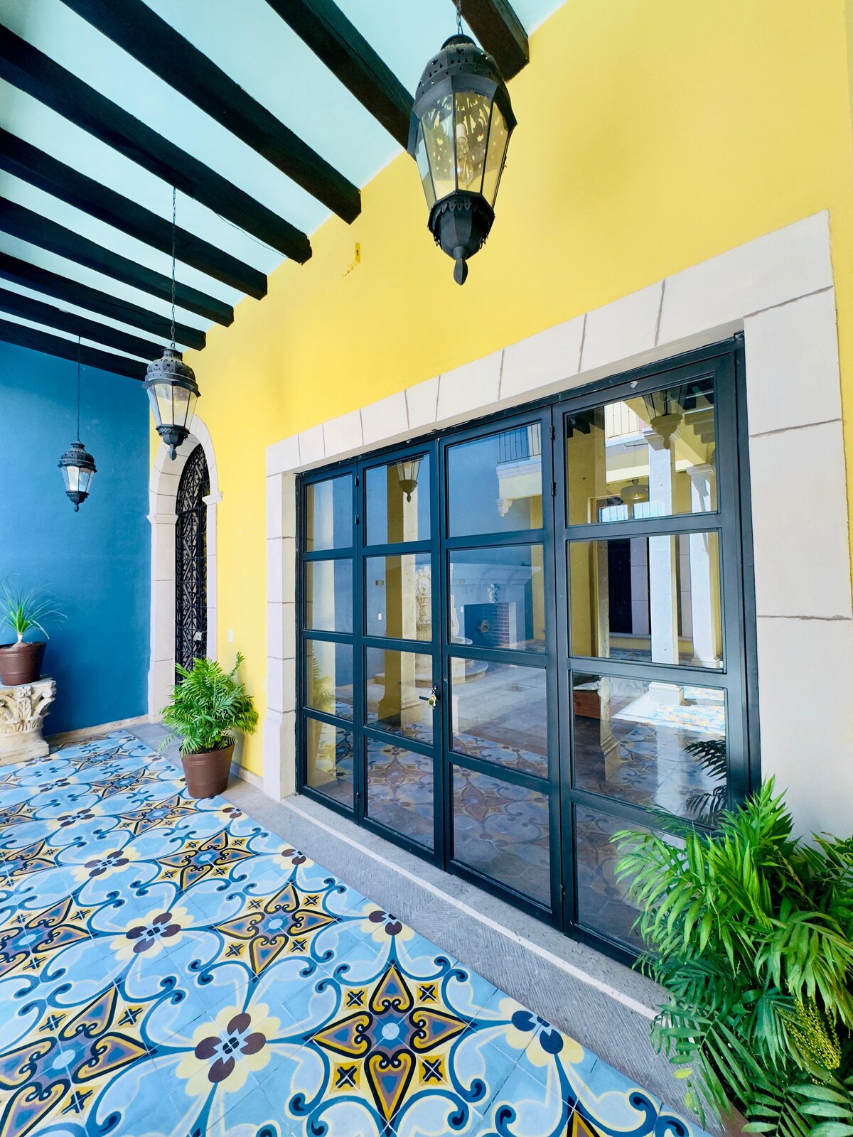 A vibrant corridor features a large window framed in black, revealing a view of the interior. The floor is adorned with colorful tiles, and hanging lanterns add a touch of elegance. Potted plants provide greenery, enhancing the welcoming atmosphere of the space.