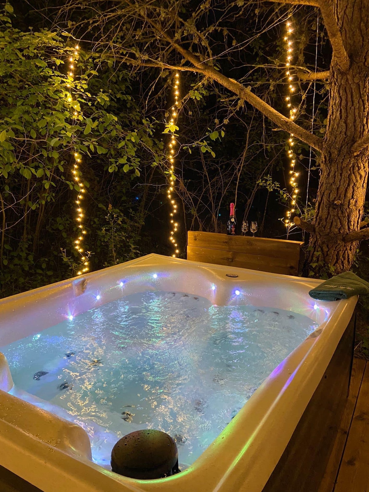 A private outdoor jacuzzi is surrounded by lush greenery and illuminated by twinkling fairy lights. The warm glow enhances the relaxing ambiance, inviting moments of tranquility. A wooden backdrop is visible, adding to the cozy setting.