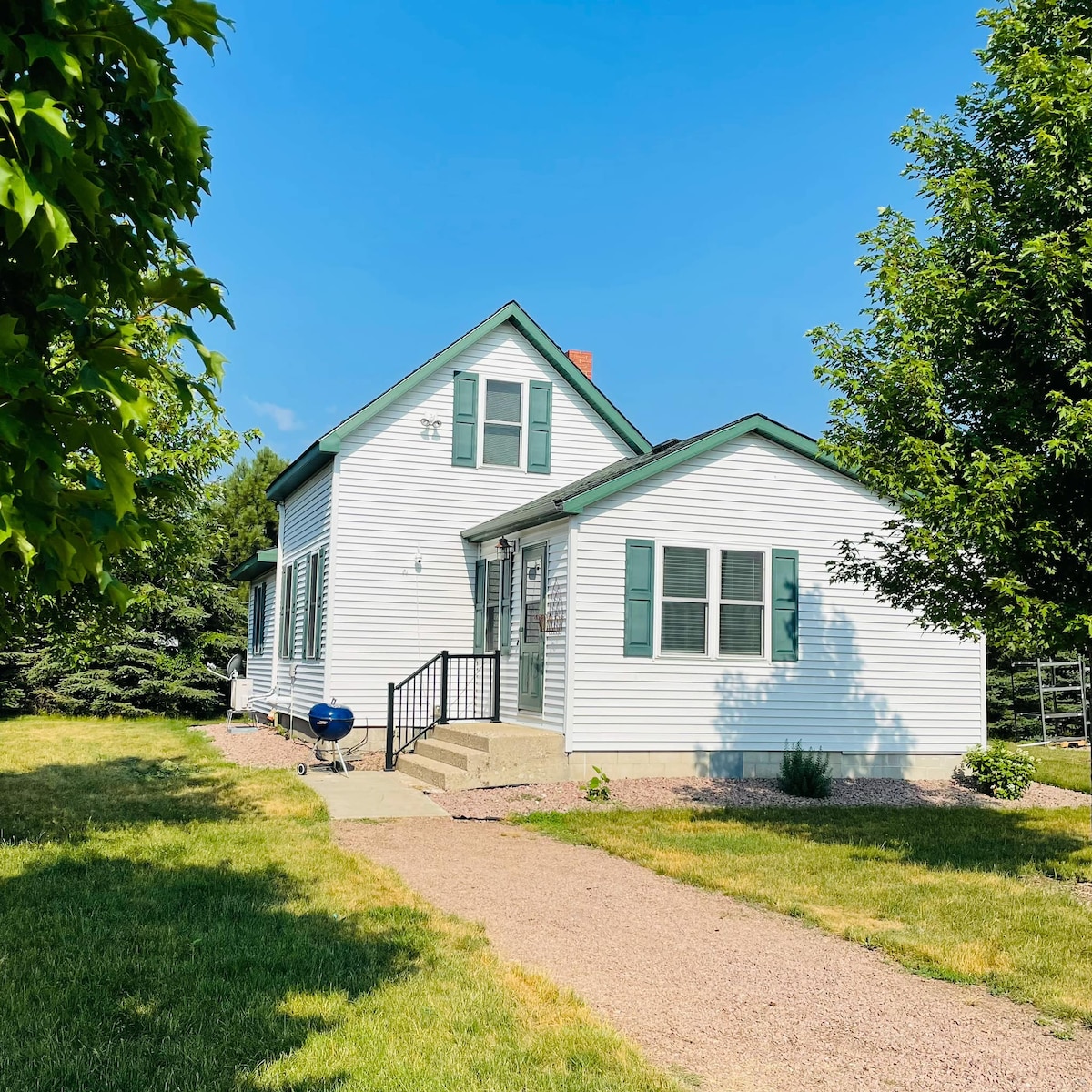 A charming two-story home with white siding overlooks a grassy area. Green shutters frame the windows, and a set of steps leads to the front door. A small grill is positioned beside the entrance, surrounded by trees in the landscape.