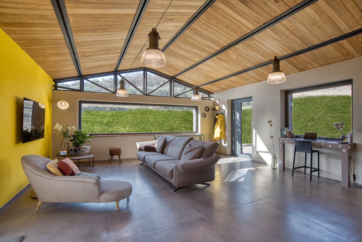 A spacious living area features a mix of modern and cozy elements, including a comfortable gray sofa and a chic accent chair. Large windows provide ample natural light, while a vibrant yellow wall adds a touch of warmth. A sleek desk stands ready for use by the window.