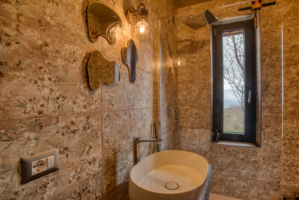 The bathroom features textured stone walls adorned with floral motifs. A modern, round sink rests atop a wooden surface. Several uniquely shaped mirrors are mounted on the wall, and a window allows natural light to enhance the space while offering a view of the outdoors.