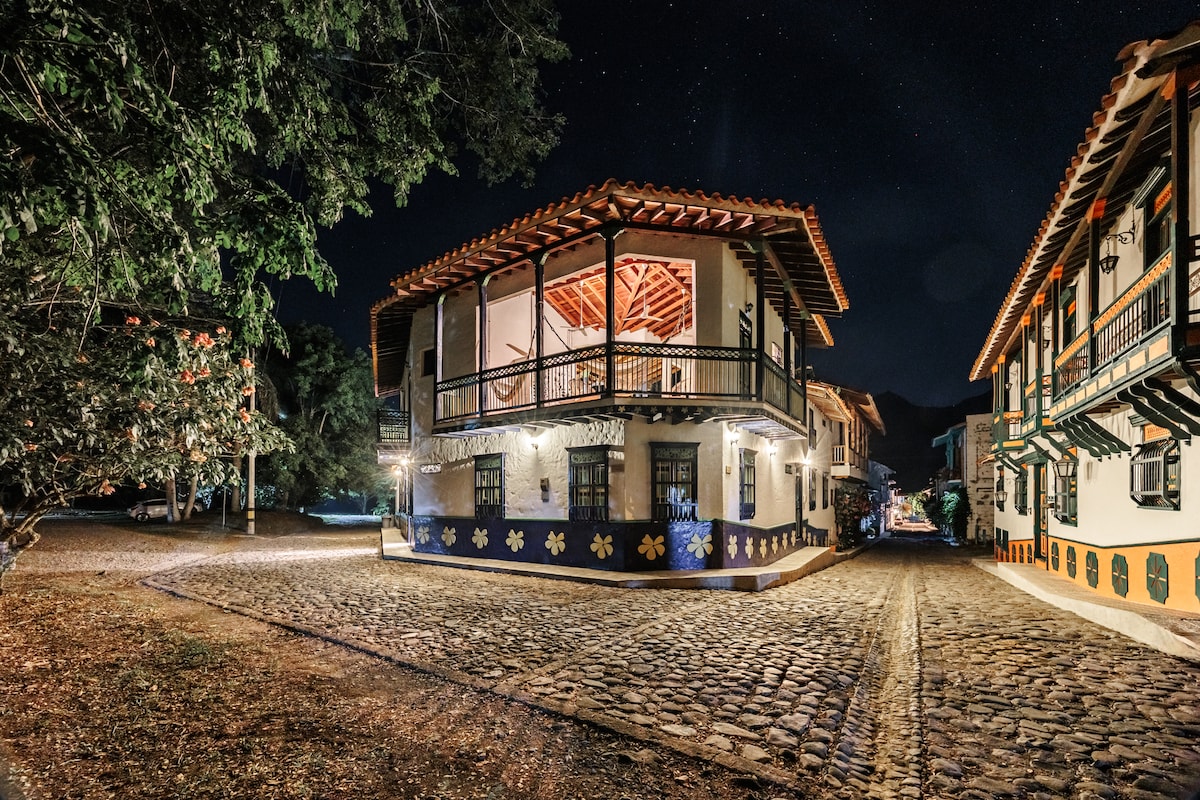 The two-story building features a traditional design with a red-tiled roof and vibrant painted accents. Elegant balconies overlook a cobblestone pathway, surrounded by trees. Soft lighting enhances the building's charm under the night sky, creating a welcoming atmosphere.
