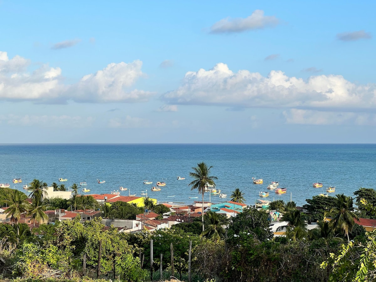 An expansive view of the coastline features a calm sea dotted with fishing boats. A backdrop of palm trees and colorful rooftops adds charm to the scene, while the horizon meets soft clouds under a clear sky.