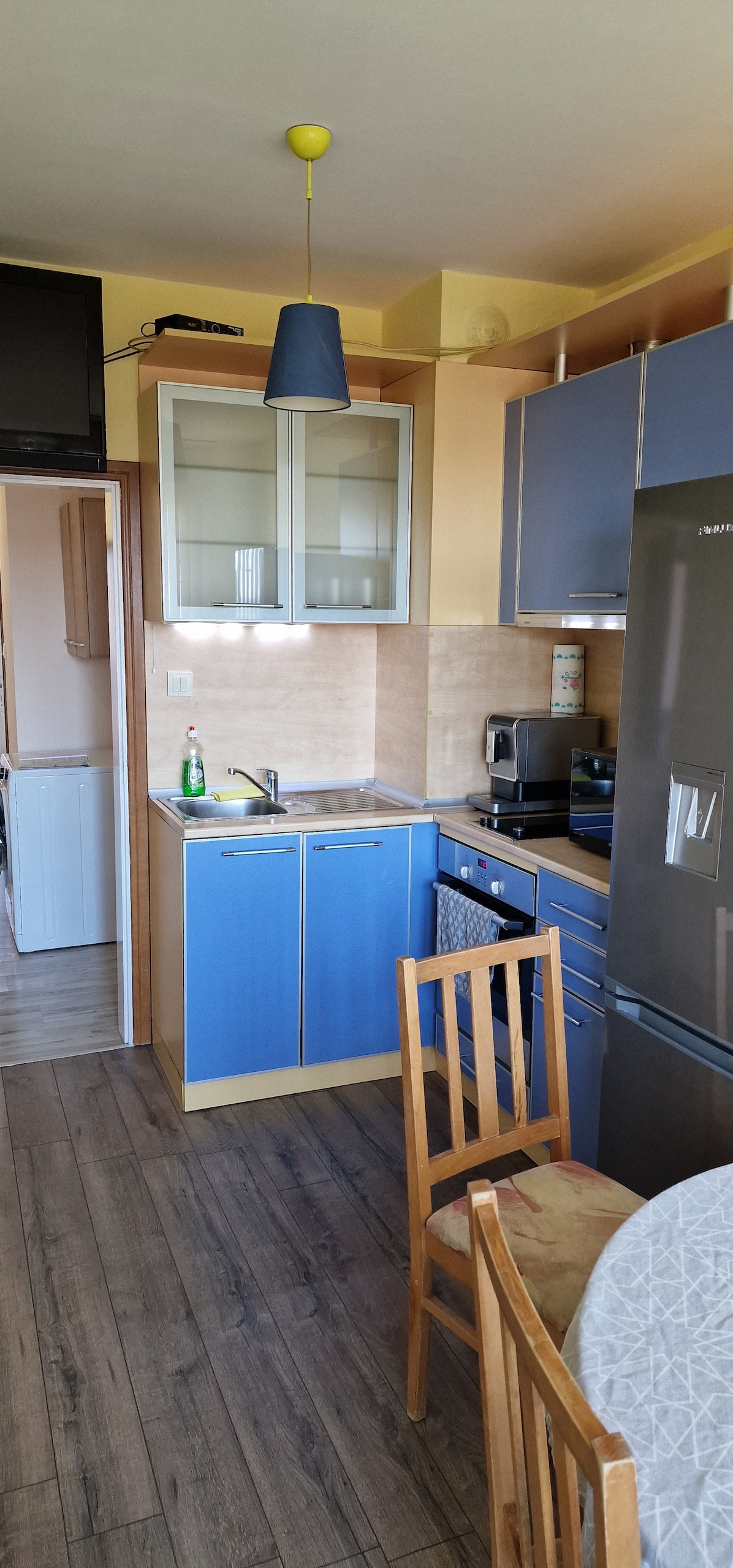 A compact kitchen area is visible, featuring blue and neutral cabinetry. Essential appliances, including a refrigerator and stove, are arranged for convenience. A small dining table with wooden chairs is positioned close by, and ample natural light brightens the space.