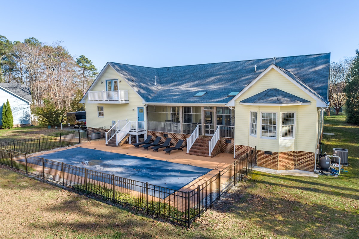 A spacious outdoor area is shown with a fenced swimming pool covered for safety. Several lounge chairs are positioned around the pool deck. The house features a light-colored exterior with multiple entries, including a set of stairs leading to a balcony.