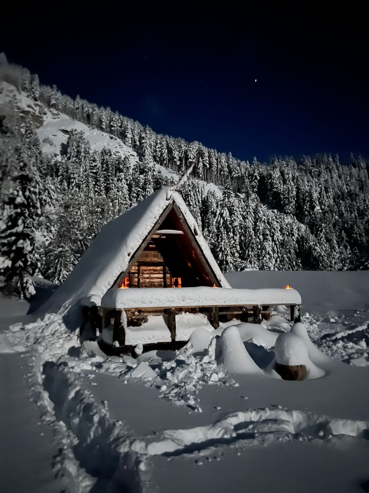 A rustic mountain cabin is surrounded by a blanket of snow, with a peaked roof and warm light glowing from within. Tall, snow-covered trees create a serene backdrop against the dark night sky, emphasizing the cabin's secluded charm.