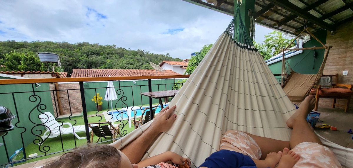 A spacious balcony area features hammocks set against a backdrop of lush greenery. A view of the pool and lawn is visible, with outdoor furniture, including tables and chairs. The setting offers a peaceful retreat, inviting relaxation outdoors.