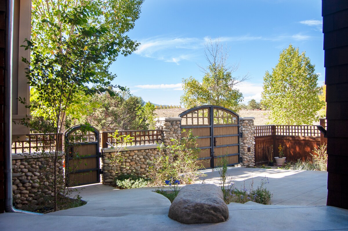 An inviting outdoor area features a stone pathway leading to dual wooden gates. Surrounding the space are lush trees and shrubs, creating a serene atmosphere. A natural stone accent can be seen on the ground, adding to the rustic charm of the entrance.