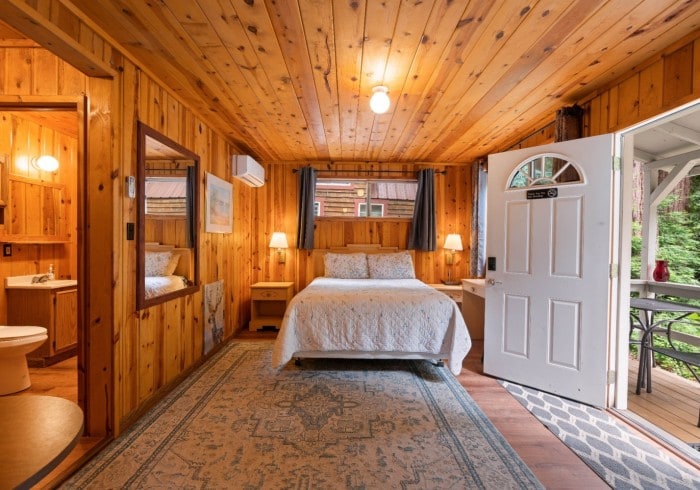 A wooden-paneled room features a spacious bed with a light-colored quilt. Soft lighting is provided by two bedside lamps. A private bathroom is visible, along with an entry door leading to an outdoor seating area. A decorative rug adds warmth to the wood floors.