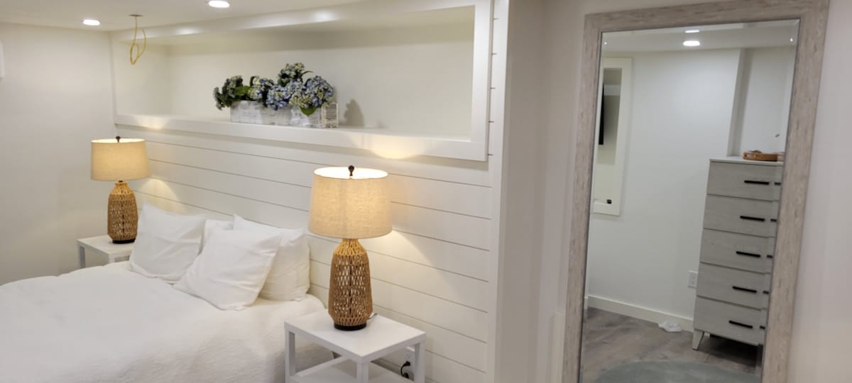 A serene bedroom setup features a spacious bed with white bedding, accompanied by two woven lamps on nightstands. Above the bed, a white shelf is adorned with decorative flowers. A full-length mirror reflects the light, enhancing the room's clean and modern design.